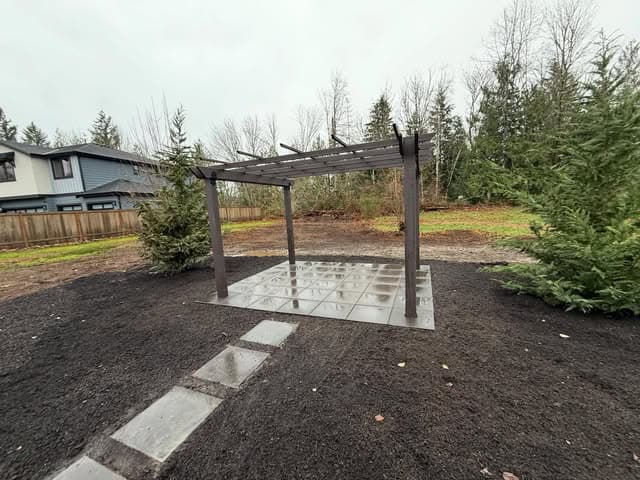 A dark-stained pergola on a paver pad, surrounded by freshly spread mulch beds and young evergreens.